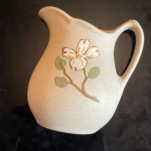 May include: A small, off-white ceramic pitcher with a handle and spout. The pitcher is decorated with a floral design featuring a white flower, green leaves, and a brown stem. The pitcher is likely used for serving liquids.
