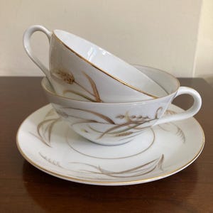 May include: Two white teacups with gold trim and wheat stalk designs are stacked on a matching saucer. The teacups have curved handles. The saucer and cups are on a dark brown surface.
