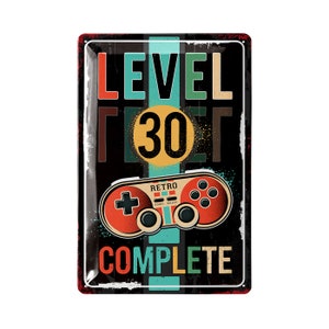 May include: A rectangular metal sign with a retro video game theme. The sign features the words "LEVEL 30 COMPLETE" in various colors, along with a vintage game controller graphic. The background is black.