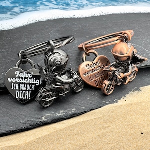 Op de afbeelding: Twee metalen motorfietsspelden. Eén is zwart, de andere is koperkleurig. Elke speld heeft een stripfiguur op een motorfiets, met een hartvormige bedel met de tekst "Fahr vorsichtig! ICH BRAUCH DICH!"