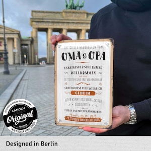 Pu&ograve; includere: Un cartello metallico rettangolare con testo tedesco, tra cui "OMA & OPA" e "OFFIZIELLE HAUSREGELN". Il cartello ha un aspetto vintage e presenta regole familiari. Lo sfondo mostra la Porta di Brandeburgo a Berlino.