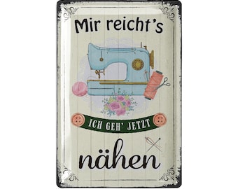 May include: A rectangular metal sign with a distressed finish features a vintage sewing machine in teal. The sign includes text in German, a pink yarn ball, a spool of thread, scissors, and floral accents, evoking a retro craft theme.