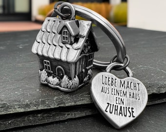 May include: A silver-colored house-shaped keychain with a heart-shaped tag. The house has a detailed roof, windows, and door. The heart tag has the German text "LIEBE MACHT AUS EINEM HAUS EIN ZUHAUSE" engraved on it.