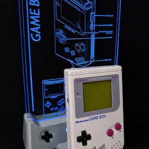 Peut inclure: Une console de jeu portable gris clair avec un écran vert, des boutons A et B violets et une croix directionnelle noire, posée sur un support assorti. Derrière se trouve un panneau en acrylique transparent avec un schéma bleu lumineux.