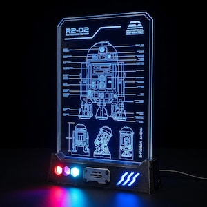 May include: A rectangular acrylic light display featuring a blue laser-etched technical schematic of the R2-D2 droid. The base is dark grey with three illuminated buttons in red, purple, and blue, and a blue light strip on the right side.