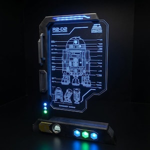 Może przedstawiać: Prostokątny, podświetlany stojak ekspozycyjny z niebieskim, świecącym schematem technicznym droida astromechanicznego R2-D2. Rama jest ciemnoszara z wbudowanymi zielonymi i niebieskimi diodami LED, prezentującymi etykiety techniczne.