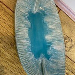 May include: An elongated, boat-shaped ceramic dish featuring a vibrant turquoise centre that transitions into a textured, light blue and white rim. The surface displays a distinct radial pattern of fine, etched lines radiating outward from the centre.