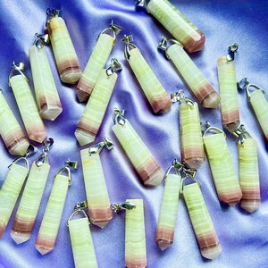 May include: A collection of gemstone pendants with silver-colored bails. The pendants are rectangular with a faceted, pointed bottom. The stones have layers of pale green, white, and mauve colors. The pendants are arranged on a purple satin fabric.