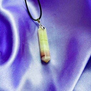 May include: A pendant necklace featuring a multi-colored, rectangular crystal. The crystal has bands of green, white, and maroon. The pendant hangs from a black cord with a silver-colored bail. The background is a purple satin fabric.