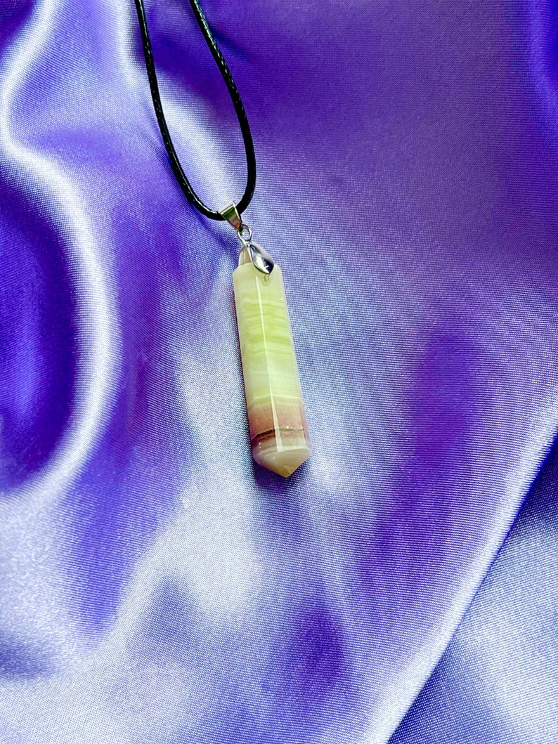 May include: A pendant necklace featuring a multi-colored, rectangular crystal. The crystal has bands of green, white, and maroon. The pendant hangs from a black cord with a silver-colored bail. The background is a purple satin fabric.