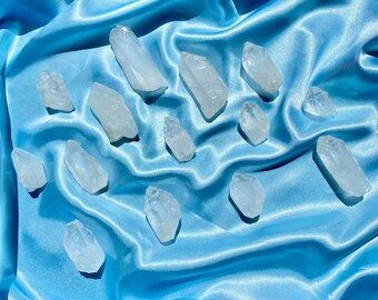 Peut inclure: Plusieurs pointes de cristal de quartz transparentes de tailles variées, disposées sur un tissu de satin bleu clair. Les cristaux ont des formes naturelles irrégulières et sont translucides.