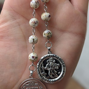 May include: A silver rosary with white and red speckled beads and two silver charms. One charm features a depiction of Saint Christopher and the text "Saint Christopher Protect Us". The other charm features a prayer in a wreath.