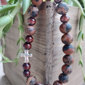 May include: A brown and black beaded bracelet with a silver cross charm. The beads are made of natural stone and have a speckled pattern.