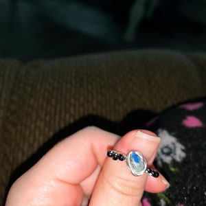 Op de afbeelding: Een zilveren draadgewikkelde ring met een druppelvormige, blauw-witte steen. De ring heeft kleine zwarte kralen aan weerszijden van de steen. Een uniek en elegant sieraad.
