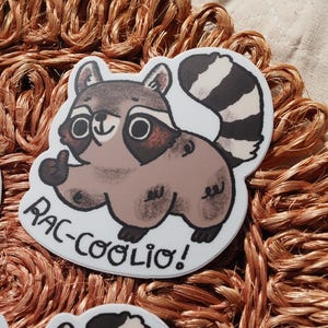 May include: A white sticker with a cartoon raccoon giving a thumbs up. The raccoon is brown and gray with a black and white striped tail. The text "Rac-Coolio!" is written below the raccoon.