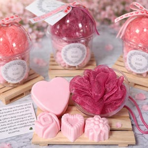 Puede incluir: Exfoliantes de azúcar y jabones rosas en forma de corazón y rosa. Los exfoliantes están en vasos transparentes con lazos rosas, y los jabones son de color rosa claro. Los productos se exhiben en pequeñas paletas de madera.