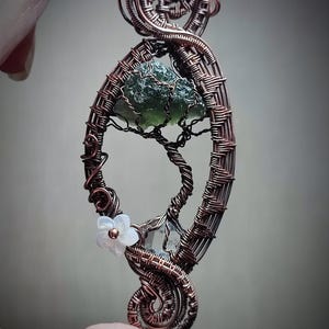 Puede incluir: Colgante artesanal de alambre de cobre con una piedra preciosa verde y un diseño de árbol de alambre. El colgante ovalado está adornado con una pequeña flor blanca. El intrincado trabajo de alambre crea una joya artística única.