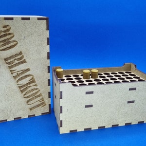 May include: A wooden box with a lid that says "300 BLKOUT" on the front. The box has a grid of holes on the top and is filled with small, brown, cylindrical objects.
