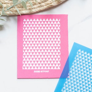 May include: A pink stencil with a white triangle pattern and the text "ECHO STONE" printed on the bottom. The stencil is on a white surface next to a blue stencil with the same pattern.