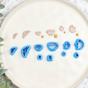 Puede incluir: Una colección de cortadores de galletas con forma de cruasanes, tazas de té, teteras y otras formas. Los cortadores son azules y beige, dispuestos sobre un plato blanco con un borde azul claro. Una ramita de hojas verdes está en el fondo.