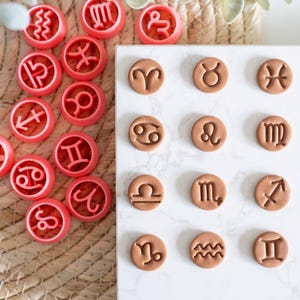 Può includere: Una collezione di timbri con simboli zodiacali rosa e marroni. I timbri rosa sono disposti su una superficie tessuta, mentre i timbri marroni sono su una superficie bianca. I timbri presentano vari segni astrologici.