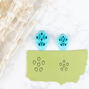 May include: Two blue plastic flower-shaped cookie cutters, one larger than the other, are shown on a light green surface. The cutters have been used to create impressions in the surface, which are visible in the image.