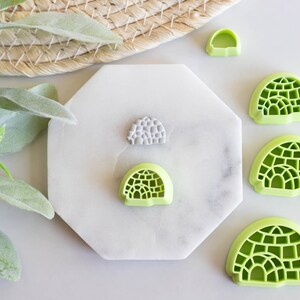 May include: Five green plastic cookie cutters shaped like igloos, with one gray plastic cookie cutter shaped like an igloo in the center. The cookie cutters are on a white marble surface.