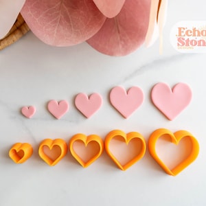 May include: A set of pink heart-shaped fondant cutters and orange heart-shaped cookie cutters of various sizes. The cutters are arranged in order of size, with the smallest cutter on the left and the largest cutter on the right. The cutters are on a white marble surface. Echo Stone Designs is printed on a yellow sun with a white background.
