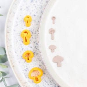 May include: A set of five yellow mushroom-shaped cookie cutters and five brown mushroom-shaped cookie cutters on a white plate with a blue and white speckled pattern.