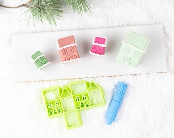 May include: A collection of miniature gingerbread house decorations in shades of pink, green, and white, with white icing details. A green house-shaped cookie cutter and a blue textured rolling pin are also present, suggesting a baking theme.