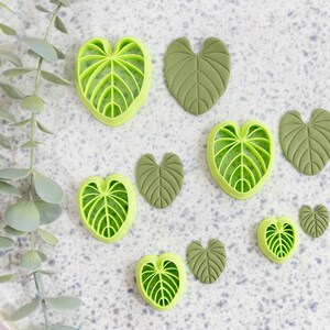 Puede incluir: Un conjunto de nueve cortadores de galletas con forma de hoja verde sobre una superficie blanca moteada. Los cortadores son de varios tamaños y formas, algunos tienen un patrón de vetas más detallado que otros.