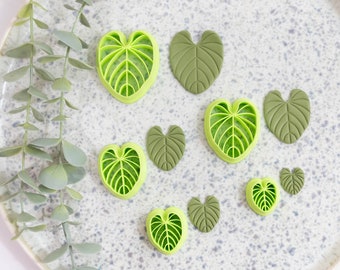 Puede incluir: Un conjunto de nueve cortadores de galletas con forma de hoja verde sobre una superficie blanca moteada. Los cortadores son de varios tamaños y formas, algunos tienen un patrón de vetas más detallado que otros.