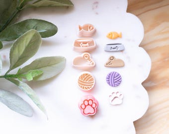 May include: A collection of cat-themed cookie cutters in various shapes, including fish, food bowls, a mouse, a ball of yarn, a paw print, and a cat-shaped bowl. The cutters are in shades of pink, yellow, beige, and purple.