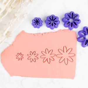 May include: A set of four purple plastic flower cookie cutters of different sizes, with the largest cutter being used to cut out a flower shape from a sheet of pink fondant.