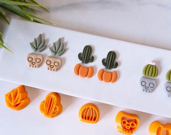 May include: A set of six pairs of polymer clay earrings featuring various Halloween and autumn-themed designs, including pumpkins, cacti, and skulls. The earrings are displayed on a white surface.