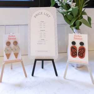 Könnte beinhalten: Zwei Paar Ohrringe, die auf kleinen Staffeleien ausgestellt sind. Das erste Paar hat ein geometrisches Design mit beige und braunen Akzenten. Das zweite Paar hat ein Schmetterlingsdesign mit orange und schwarzen Akzenten. Beide Sets sind mit "Echo Stone Jewelry" gekennzeichnet.