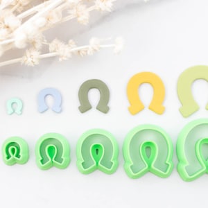 May include: Set of nine green, blue, and yellow plastic cookie cutters shaped like horseshoes. The cutters are arranged in order of size, with the smallest cutter on the left and the largest cutter on the right.