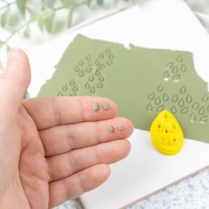 May include: A hand holding several small green clay leaf shapes. A green clay sheet with a pattern of leaf shapes is in the background. A yellow plastic leaf mold is on the sheet.