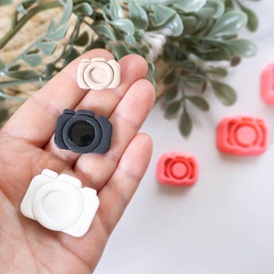 May include: Five miniature camera shaped charms in different colours. The charms are in a hand, with three of the charms in the palm of the hand and two on the table.