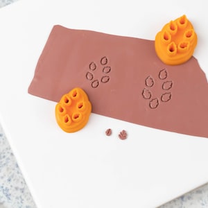 May include: A close-up of a brown clay surface with orange leaf-shaped stamps. The stamps have been pressed into the clay, creating a pattern of leaves. There are also two smaller orange leaf stamps on the white surface.