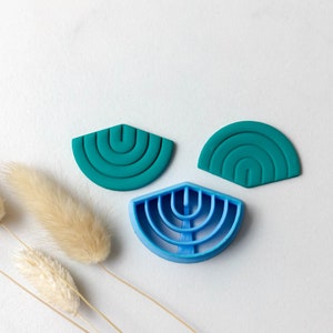 Peut inclure: Trois emporte-pièces en silicone en forme de demi-cercle avec un motif arc-en-ciel. Les emporte-pièces sont turquoise, turquoise et bleu.