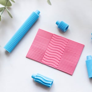 Puede incluir: Una hoja texturizada rosa con un patrón ondulado en el centro. Cuatro aplicadores de plástico azul con texturas acanaladas están dispersos alrededor de la hoja.