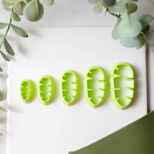 May include: Five green plastic leaf-shaped cookie cutters of varying sizes. The cutters are arranged in a row from smallest to largest.
