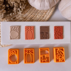 Puede incluir: Conjunto de seis sellos de arcilla polimérica naranja y marrón con diseños otoñales. Los sellos tienen forma de rectángulo y presentan una bellota, una calabaza, un hongo y una hoja.