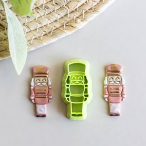 May include: Three nutcracker cookie cutters, two are pink with gold trim and one is green. The green cutter is in the centre and the pink cutters are on either side.