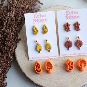 May include: Two white display cards labeled Echo Stone Designs featuring yellow and brown leaf and tree earrings. Below are four orange plastic cutters in leaf and tree shapes. The yellow earrings include studs and dangles with gold-tone posts.