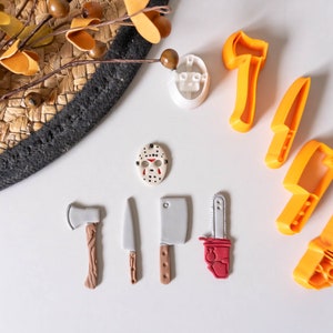 May include: A set of orange plastic Halloween-themed cookie cutters shaped like an axe, knife, machete, and chainsaw. There is also a white plastic cookie cutter shaped like a hockey mask and a small red and white plastic cookie cutter shaped like a chainsaw.