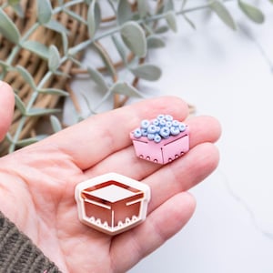 Puede incluir: Una mano sostiene dos pequeños objetos decorativos. Uno es una caja rosa llena de esferas azules, que parecen arándanos. El otro es una forma de caja geométrica, abierta por arriba, con un acabado en oro rosa. Probablemente son suministros para manualidades o componentes de joyería.