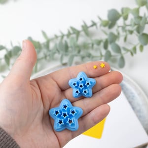May include: Two blue star-shaped cookie cutters with a star-shaped design on each. The cookie cutters are on a hand against a white background.