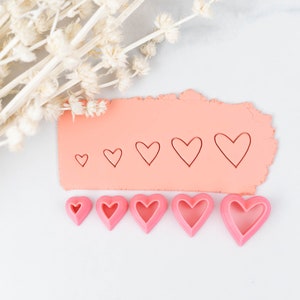 May include: A set of six pink heart-shaped cookie cutters of varying sizes. The cutters are arranged in a row on a white surface, with a pink clay surface behind them. The clay surface has five heart shapes cut out of it, also in varying sizes.